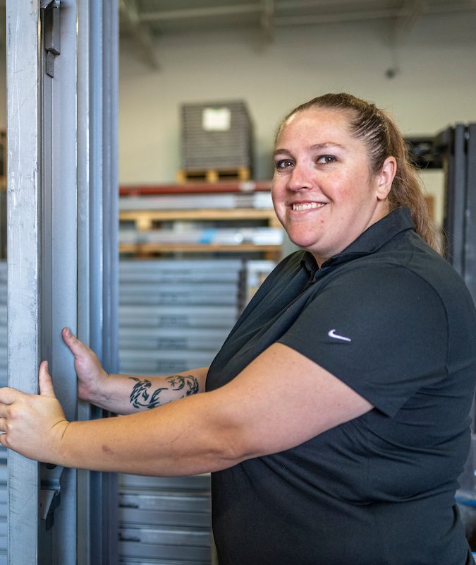 Employée de Raymond Architectural souriante manipulant des cadres architecturaux en acier dans l’atelier – Raymond Architectural