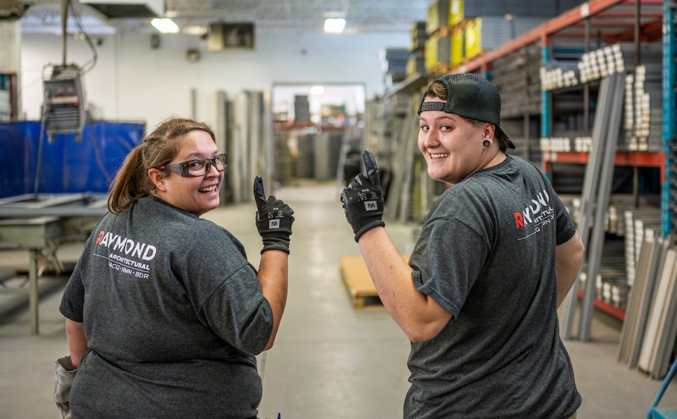 Deux employés souriants de Raymond Architectural dans l’atelier de fabrication de portes et cadres en acier, illustrant l’esprit d’équipe – Raymond Architectural