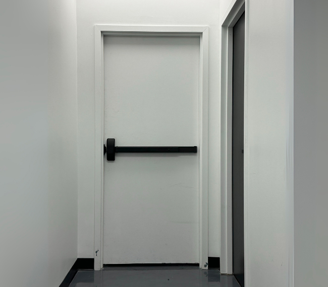 Porte coupe-feu blanche en acier avec barre anti-panique installée dans un couloir institutionnel – Raymond Architectural