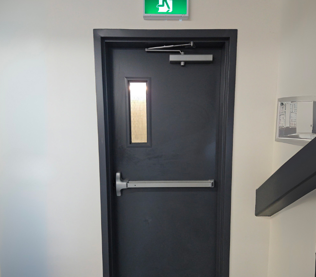 Porte coupe-feu en acier gris avec barre anti-panique et vitrage étroit, installée comme sortie de secours – Raymond Architectural